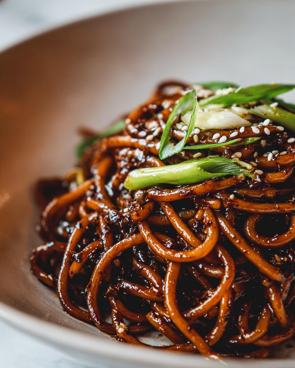 A close-up shot of glossy, dark brown Asian garlic noodles topped with sliced green onions and sesame seeds.