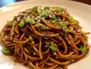 A close-up shot of a serving of glossy, dark brown Asian garlic noodles topped with sliced green onions and sesame seeds.