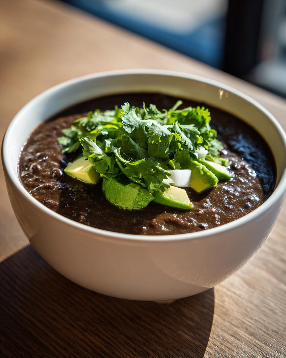 A close-up of a white bowl filled with thick black bean soup, topped with fresh avocado slices and cilantro.