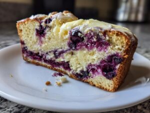 A thick slice of moist Blueberry Cream Cheese Bread showing swirls of purple blueberries and cream cheese filling, topped with a light glaze.