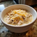 A close-up of a white bowl filled with creamy white chili, featuring shredded chicken and topped generously with mixed shredded cheddar and Monterey Jack cheese.