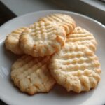 A stack of five golden, textured butter cookie treats resting on a white plate near a window.