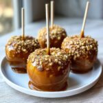 Four glossy caramel apple treats coated in chopped nuts, served on a white plate.