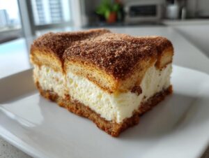 A square slice of creamy churro cheesecake topped with cinnamon sugar on a white plate.