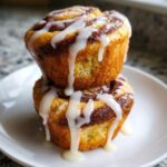 Two stacked cinnamon roll muffins drizzled generously with white vanilla icing on a white plate.
