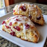 Close-up of two flaky cranberry orange scones topped with a thick white glaze, studded with dried cranberries.