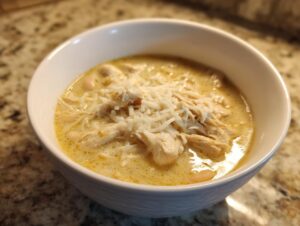 A close-up of a white bowl filled with creamy, yellow-hued cream cheese chicken chili, topped with shredded white cheese.