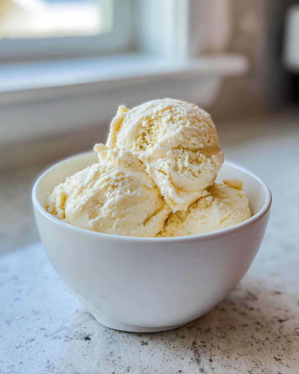 Three scoops of creamy, pale yellow banana ice cream served in a white bowl on a speckled countertop.