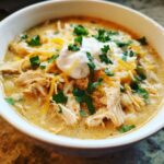 A close-up of a bowl of creamy white chicken chili, garnished with shredded cheese, sour cream, and fresh cilantro.
