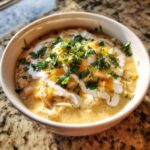 A close-up of a bowl of creamy chicken chili recipe topped with sour cream, shredded cheddar, and fresh cilantro.