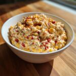 A close-up of creamy, cheesy corn dip mixed with red peppers and spices, served in a white bowl.