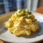 A mound of creamy, yellow egg salad recipe garnished with fresh chopped chives on a speckled white plate.