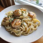 Close-up of creamy seafood pasta topped with four golden-brown seared scallops and fresh parsley.