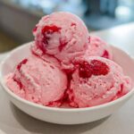 Close-up of four scoops of creamy strawberry ice cream with visible fruit swirls in a white bowl.
