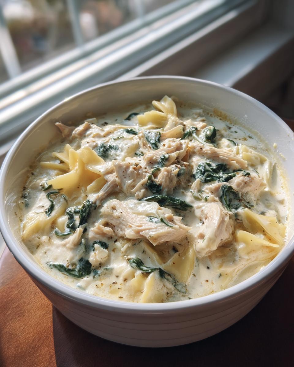 A close-up of a white bowl filled with creamy white chicken lasagna soup, featuring shredded chicken, spinach, and bowtie pasta.