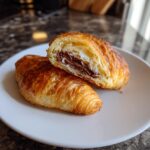 A golden brown chocolate croissant cut in half, revealing the melted chocolate filling and flaky interior.