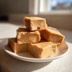 A stack of creamy, light brown maple fudge squares piled on a small white plate.