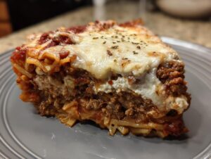 A thick, layered slice of baked spaghetti pie topped with melted mozzarella cheese and herbs.