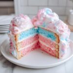 A cross-section slice of a colorful cotton candy cake showing pink and blue layers, topped with fluffy frosting.