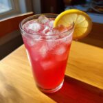 A tall glass filled with bright red pink lemonade, ice cubes, and garnished with a lemon slice.