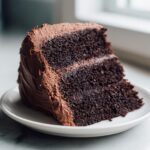 A close-up of a moist slice of homemade chocolate cake layered and covered in rich chocolate frosting.