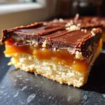A close-up, cross-section view of a homemade twix bar slice showing a shortbread base, thick caramel layer, and chocolate topping.
