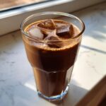 A tall glass filled with rich, dark iced chocolate milk and several large ice cubes, sitting on a marble counter.
