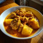 A close-up, appetizing bowl of Jamaican curry chicken served with large chunks of yellow potato in a rich, golden sauce.