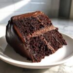 A rich, moist slice of chocolate fudge cake with thick, glossy chocolate frosting served on a white plate.