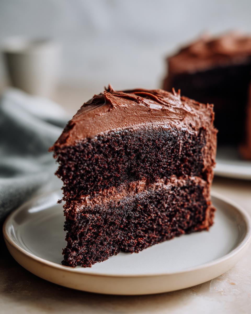 Close-up of a rich, moist slice of homemade chocolate cake with thick chocolate frosting layers.