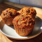 Three freshly baked morning glory muffins with visible shredded carrots sitting on a white plate in bright sunlight.