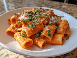 A serving of rigatoni pasta coated in creamy pink sauce pasta, garnished with shredded cheese and fresh basil.