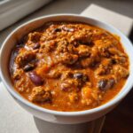 Close-up of a white bowl filled with rich, orange-red pumpkin chili containing ground meat and kidney beans.