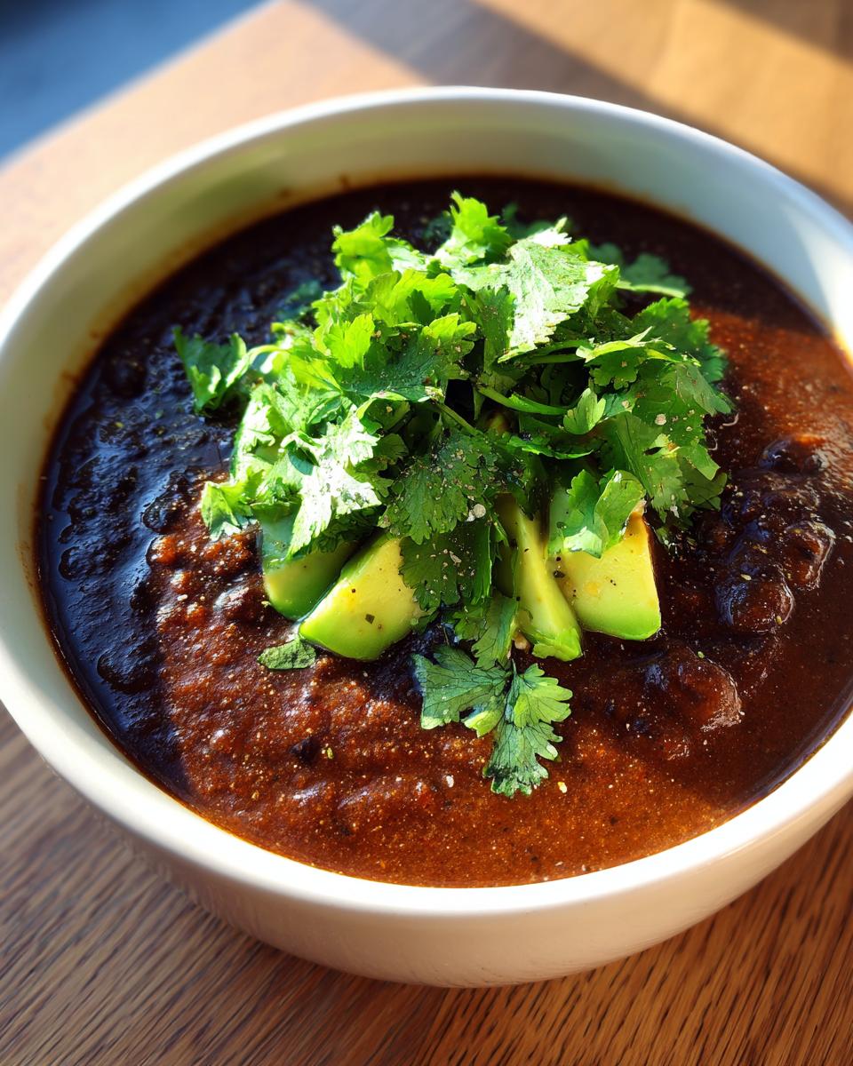 A close-up of a rich, dark bowl of black bean soup garnished with fresh avocado slices and cilantro.
