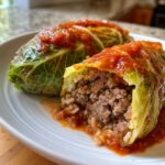 Close-up of two savory cabbage rolls topped with tomato sauce, one cut open showing the ground meat and rice filling.