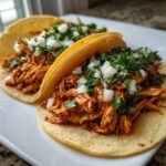 Two soft corn tortillas filled with saucy shredded chicken tacos, topped with diced white onion and cilantro.