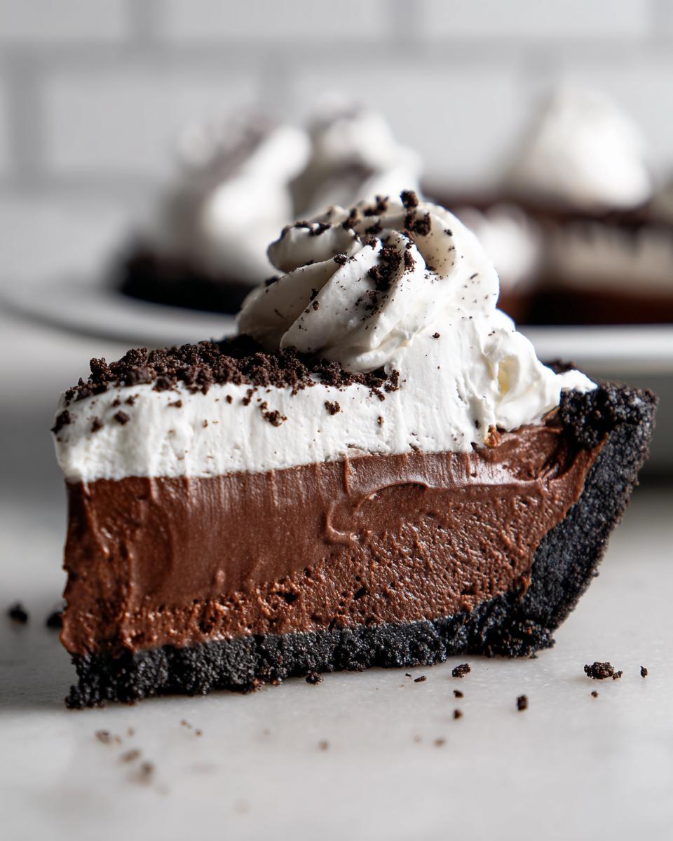 Close-up of a slice of chocolate mousse pie featuring a dark crust, two layers of chocolate filling, and a swirl of whipped cream topped with crumbs.