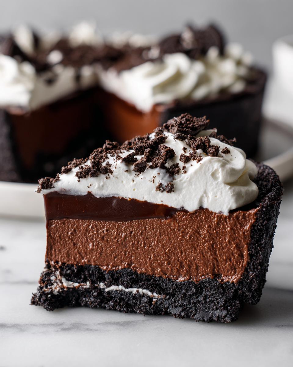 A close-up slice of decadent chocolate mousse pie featuring a dark crust, two layers of chocolate filling, whipped cream, and cookie crumbles.