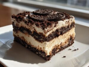 A square slice of layered dirt cake featuring alternating layers of chocolate cookie crumbs and creamy filling, topped with Oreo cookies.