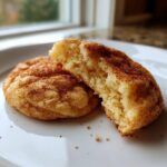 Close-up of a soft, chewy snickerdoodle cookie broken in half, showing the fluffy interior and cinnamon sugar coating.
