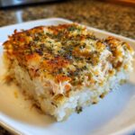 A close-up of a square serving of baked rice topped with creamy, browned crab mixture, sprinkled with furikake for the perfect sushi bake.