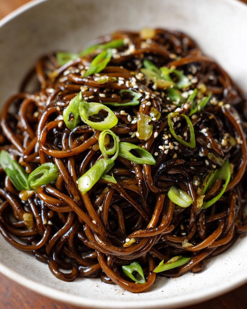 A close-up of rich, dark brown Asian garlic noodles topped with bright green scallions and sesame seeds.