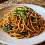 A close-up of a serving of glossy, saucy Asian garlic noodles topped with sliced green onions and sesame seeds.