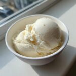 A close-up shot of creamy vanilla protein ice cream scooped into a small white bowl near a window.
