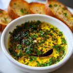 Close-up of a white bowl filled with bright yellow olive oil bread dip, topped with fresh parsley, chili flakes, and balsamic vinegar swirl.