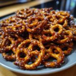A close-up shot of a pile of shiny, glazed toffee pretzels sprinkled with sesame seeds on a gray plate.