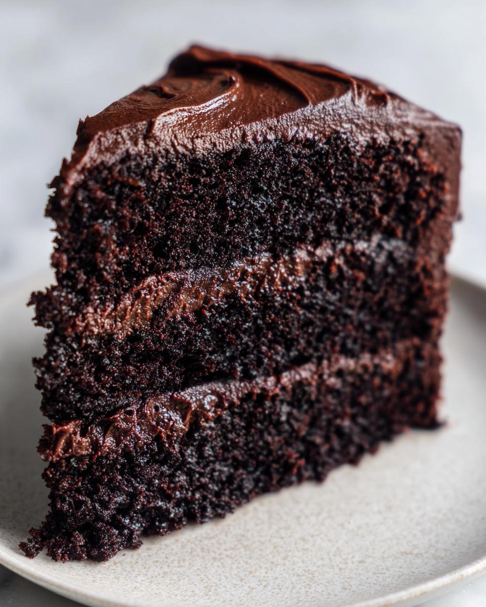 Close-up of a rich, moist slice of three-layer chocolate cake with thick chocolate frosting.