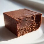 A close-up of a perfectly cut, rich, dark chocolate fudge square resting on a white plate.