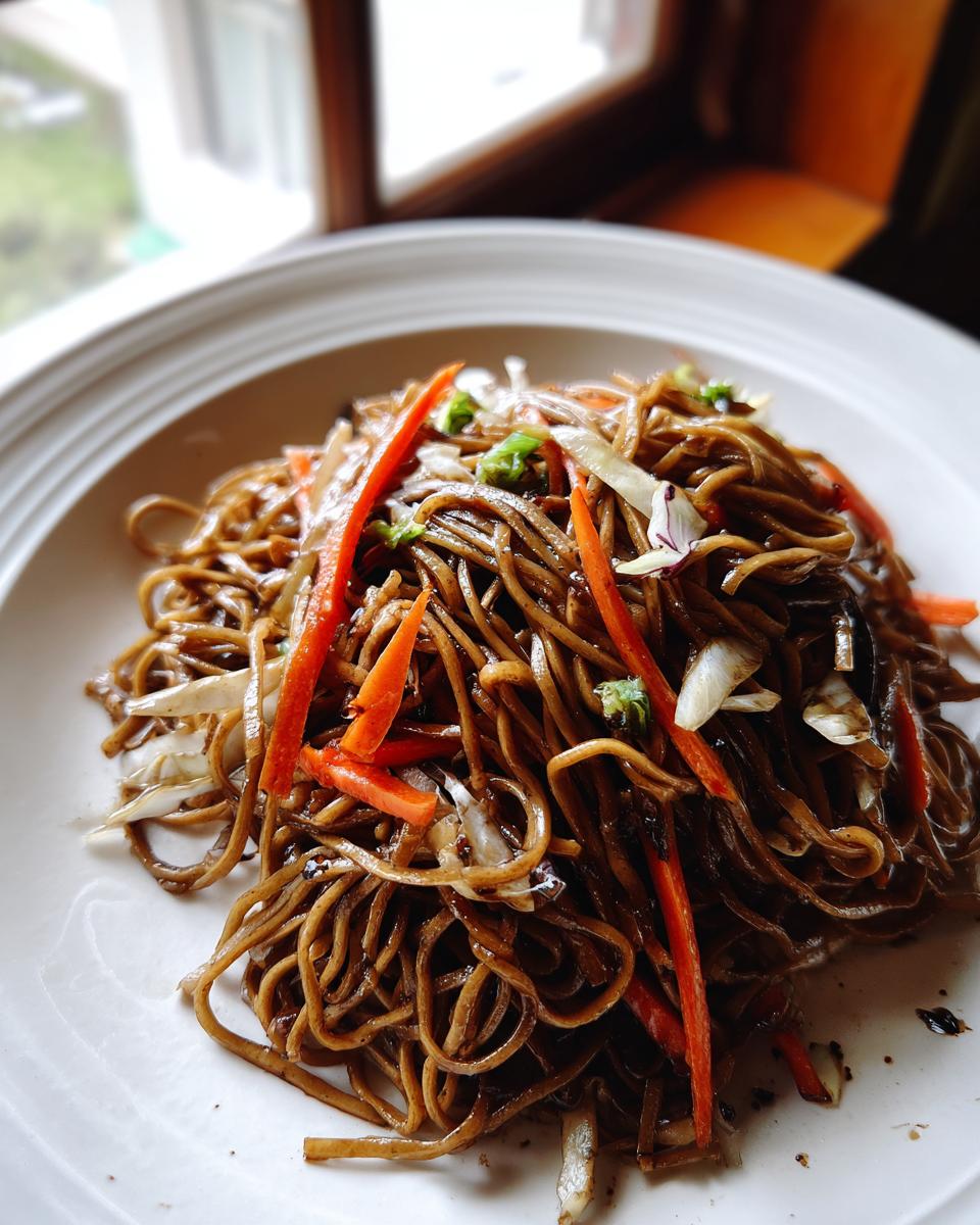 A close-up of a serving of dark, saucy fried noodles mixed with bright julienned carrots and cabbage on a white plate.