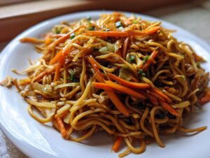 A close-up of a plate piled high with savory fried noodles mixed with julienned carrots and green onions.